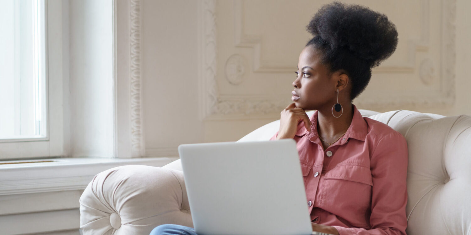 Thoughtful serious African American millennial woman with afro hairstyle wear pink shirt sitting on couch at home, distracted from laptop work looking out the window, thinking about problems.